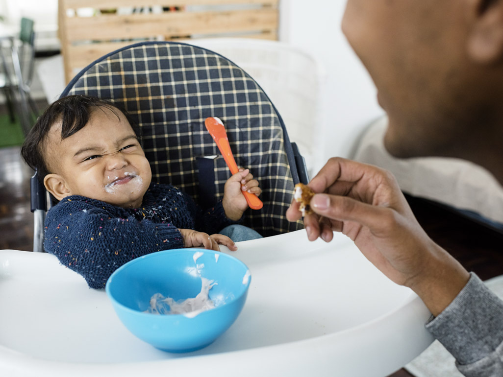baby holding orange spoon in high chair and smirking with food on his face