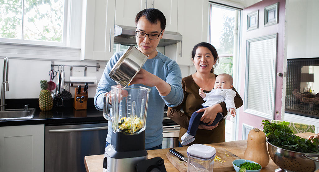 mom holding baby while looking at dad who is putting food in blender