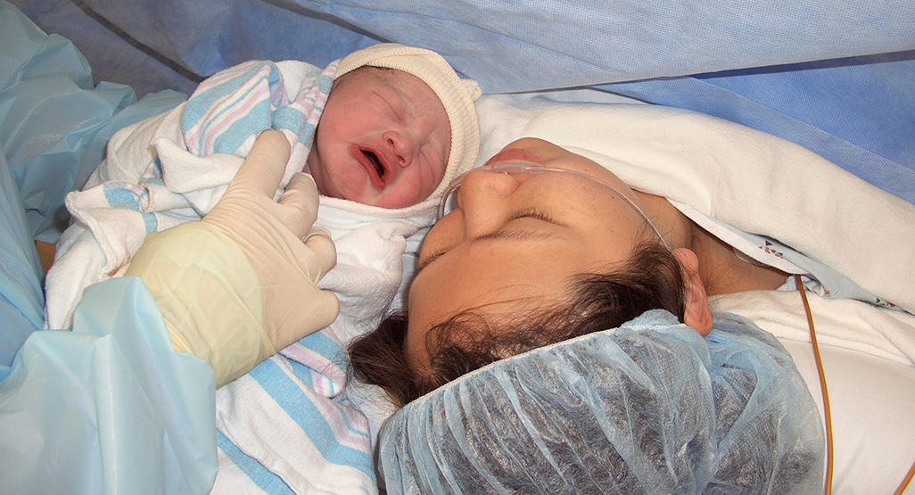 doctor showing a newborn wrapped in a towel, to a mother lying down in the operating room after a c-section