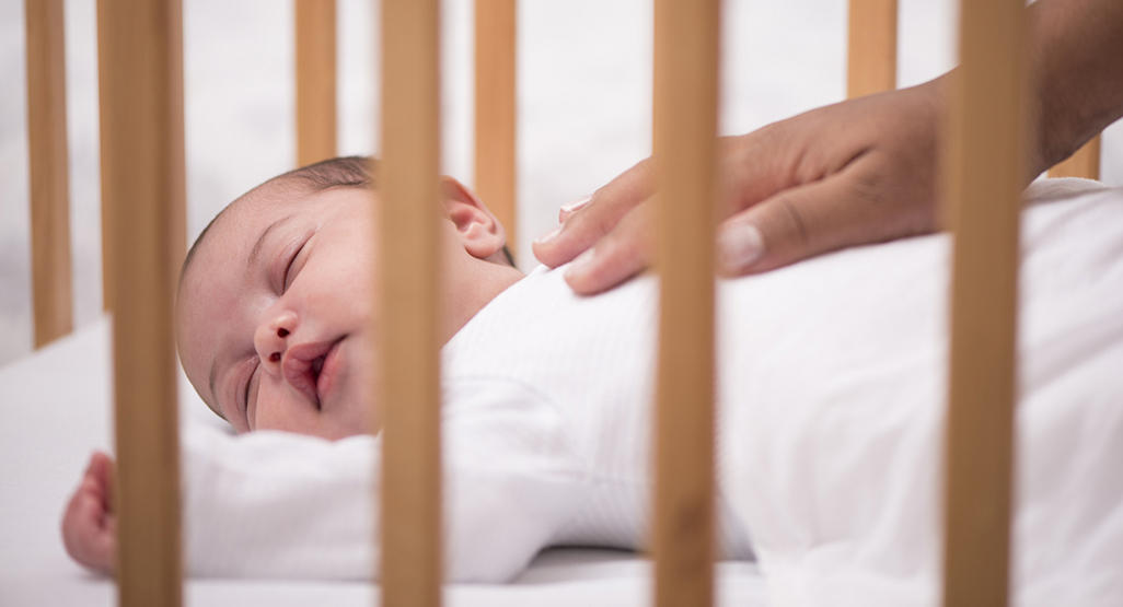 newborn sleeping on the back in his crib while the hand of grown woman is comforting him