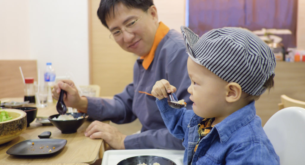 boy eating rice out of a bowl while dad looks on