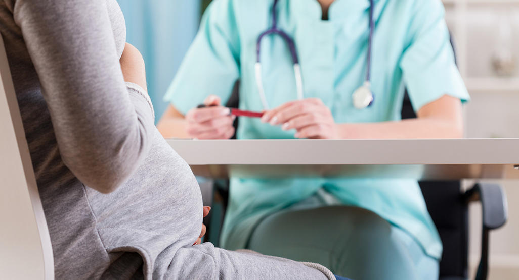 pregnant woman cradling her belly sitting across from nurse with stethoscope