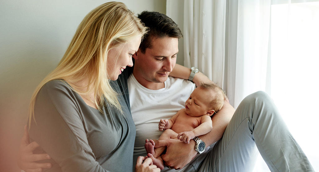 new parents sitting on a bed next to the window with their newborn in father’s arms
