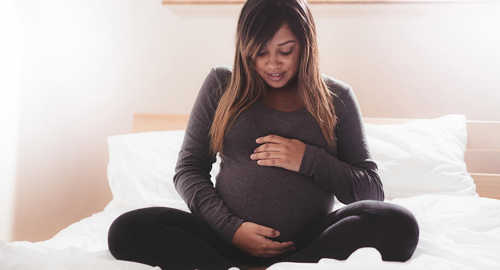 pregnant woman sitting on the bed, smiling and holding her stomach