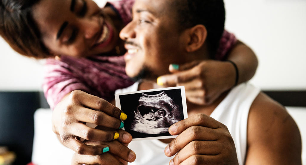 man holding an ultrasound baby photo, while a woman is hugging him from behind