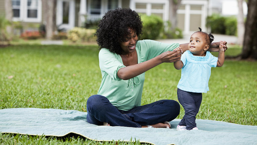 woman sitting on a blanket on grass teaching her child to walk