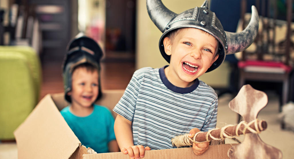 two kids in a cardboard box with viking and knight helmets on their heads, and a wooden axe in hand