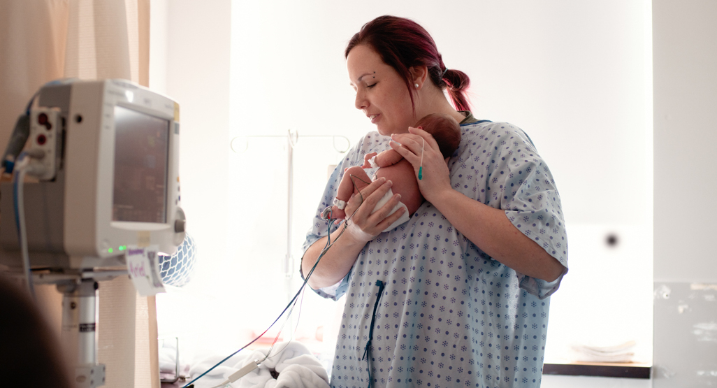 woman wearing hospital gown, holding her newborn baby in her hospital room