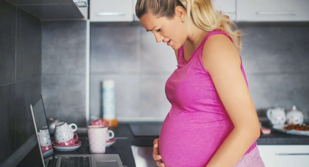 pregnant woman clutching belly while looking at laptop