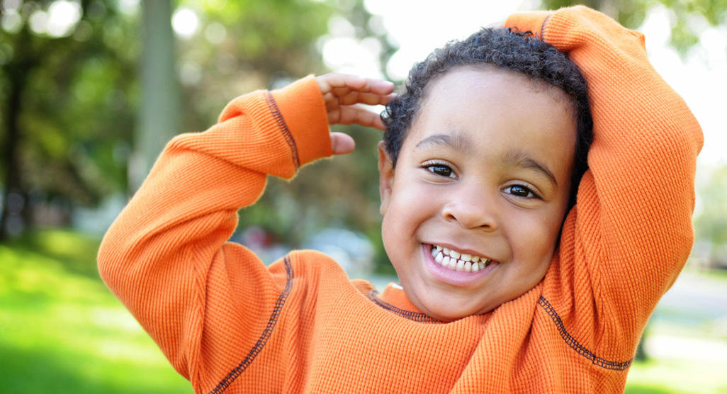 young boy in orange shirt with hands above his head smiling outdoor