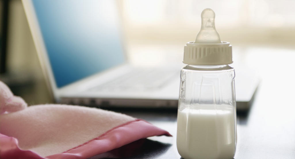 baby bottle with milk on a desk next to a laptop and a pink blanket