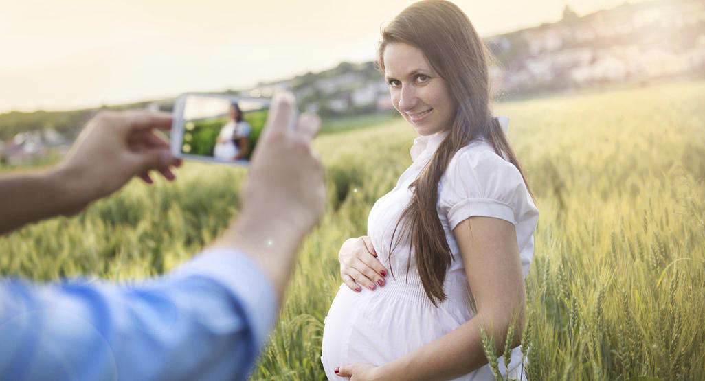 man taking a picture with his smartphone of a pregnant woman in a field of wheat