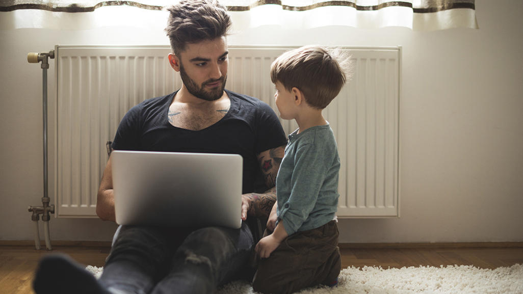 man sitting on the floor near a radiator, holding the laptop and looking at the little boy