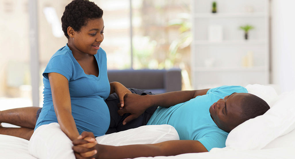 pregnant woman sitting on a bed, with her partner lying down, holding their hands