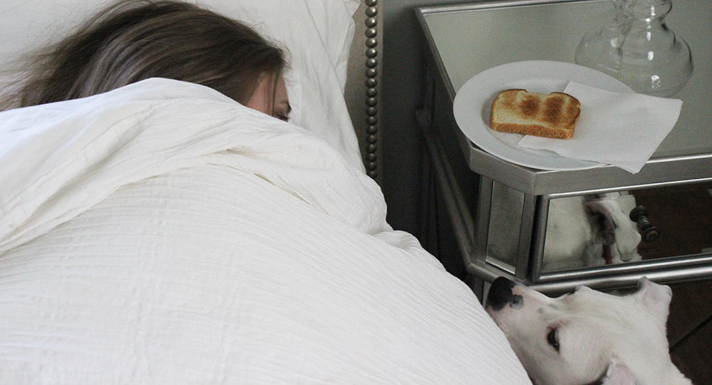 woman laying in bed under the covers with a piece of toast on the night stand next to her