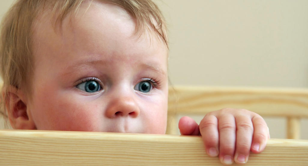baby standing up in crib while holding on to the side looking scared