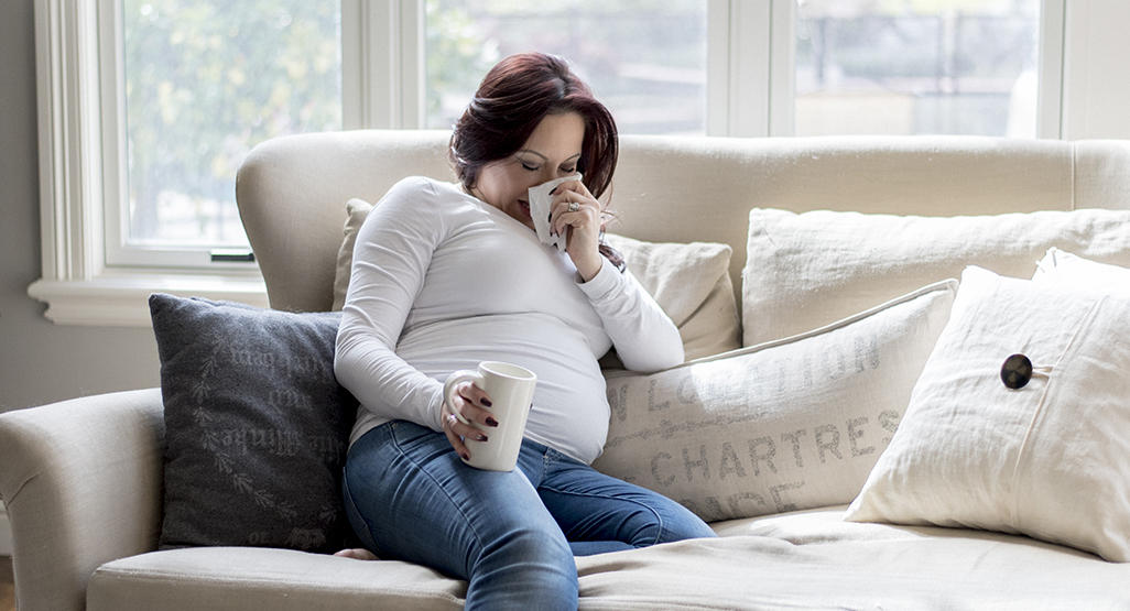 pregnant woman lying on a sofa and blowing her nose while holding a cup in her right hand