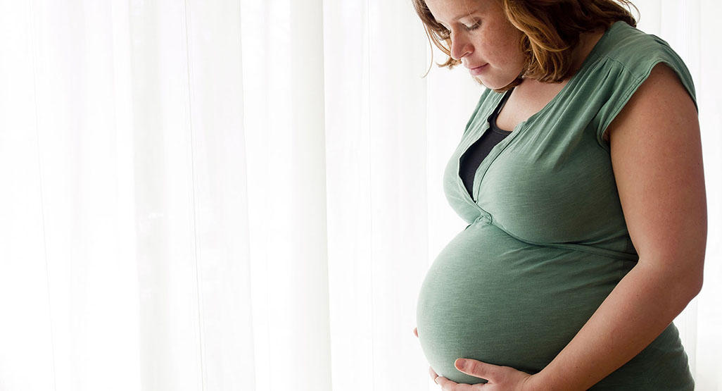 pregnant woman holding her stomach and standing next to a window with curtains