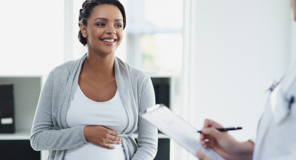 pregnant woman sitting on an exam table speaking to a physician who is making notes on a chart