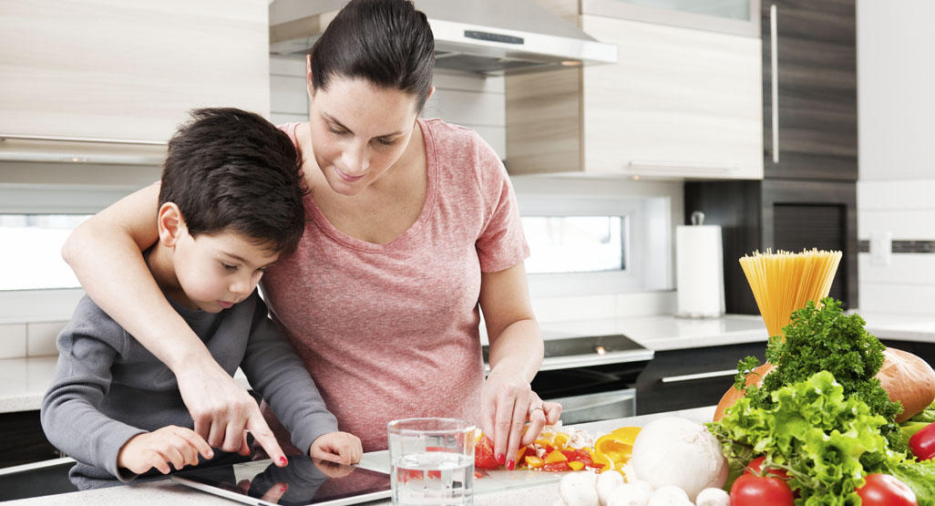 mother and son looking at a tablet on the kitchen counter, preparing vegetables for a meal