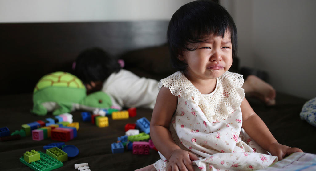 crying little girl surrounded by toys sitting on bed