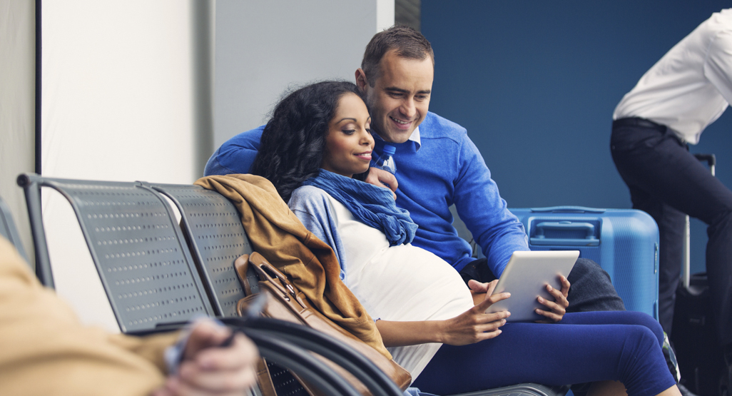 pregnant woman and her husband sitting at an airport, looking at a tablet