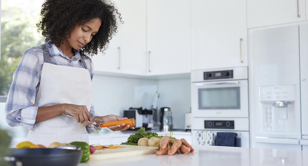 woman in the kitchen peeling carrots with a large amount of vegetables on the counter