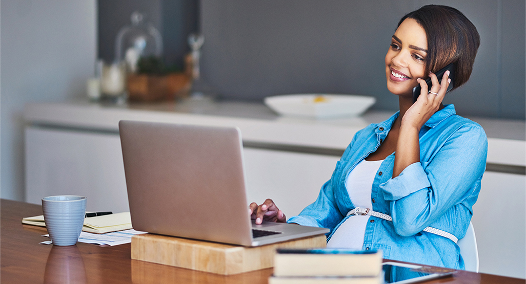 pregnant woman sitting at a table while using a laptop and talking on the phone