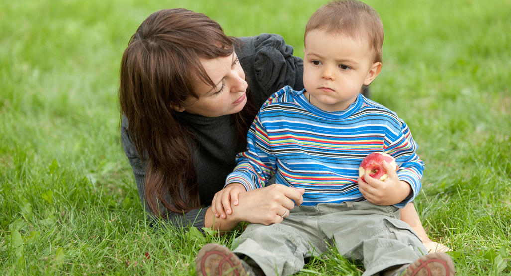 toddler and mother sitting down in a park while toddler holds an apple